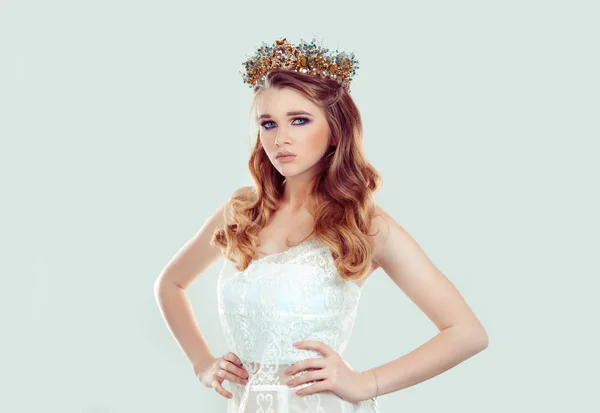 Confident beauty pageant queen. Serious woman in golden crown with blue brown stones gems crystals in white lace dress vogue style with curly wavy hair curls looking at you camera on green background