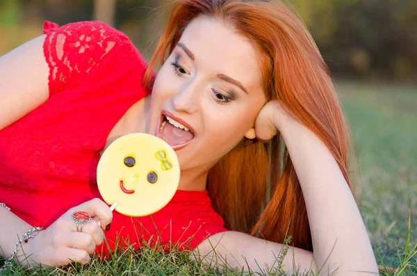 Chica feliz en un vestido rojo a punto de morder un caramelo emoticono lollipop — Foto de Stock