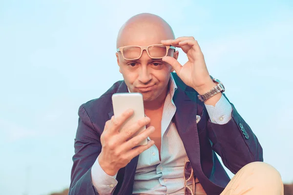 Hombre de mediana edad con gafas escéptico mirando a la pantalla del teléfono celular sentado al aire libre — Foto de Stock