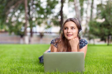Mutlu ses ders dinleme. Şehir park Üniversitesi Kampüs dışında yeşil çim çim üzerinde yalan dizüstü şarkılarla hayalifener düşünmeye ve kulaklık ile müzik dinleme Casual kız öğrenci
