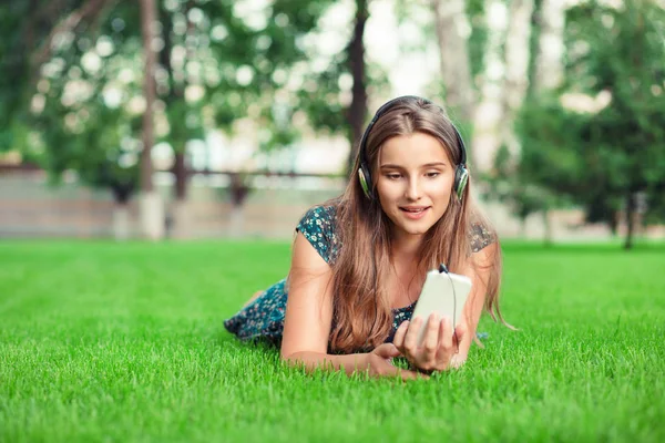 Front view of a fashion girl listening music with headphones and smart phone on line outside in park lying down on green lawn background with copy space. Multicultural model mixed race asian caucasian