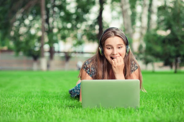 Impatient happy girl nail-biting while she waits for a web call or an answer to her chat or sms on laptop outside in park lying down on green lawn background with copy space. Multicultural  mixed race