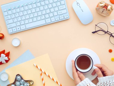 Noel kadınlarının elleri boş kağıt not, klavye ve yeni yıl süslemeleriyle dolu bir masada çay bardağını tutuyor. Kadınsı blog yazarı kış çalışma alanı. Şeftali rengi arkaplanda düz bir görünüm