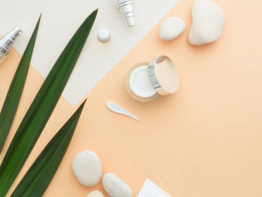 Doğal kozmetik konsepti. Taze yeşil yapraklar, yüz, göz kremi, duş jeli, sıvı sabun, bej rengi çıplak ten arka planında izole edilmiş beyaz kağıt üzerindeki spa taşları. Düz konum, üst görünüm kopyalama alanı. 