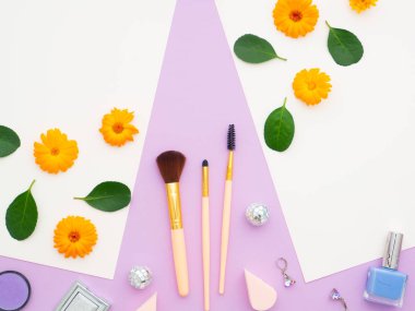 Beyaz kağıtlarda taze çiçek ve yeşil yapraklar ve doğal kozmetik ürünler, makyaj malzemeleri ve fırçalar, mor arka planda izole edilmiş ayna. Turuncu çiçeklerden yapılmış çerçeve. Düz konum, üst görünüm, kopyalama alanı