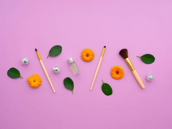 Makeup brushes, everyday make-up tools. Closeup cosmetic essentials Shampoo, green leaves, orange flowers, silver balls, on a purple background.