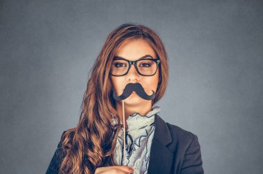 Ciddi bir erkeği taklit eden güzel, güzel, genç bir kadının portresini yakından çek. Gri arka planda sahte bıyıklı çekici bir kız. Olumlu insan duyguları, yüz ifadeleri