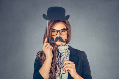 Güzel, güzel genç bir kadın bir erkeği taklit ediyor. Gri arka planda, kağıt bıyıklı ve şapkalı çekici bir kız. Portre resmini yakın çekim. Olumlu insan duyguları, yüz ifadeleri