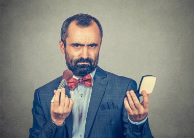 Kasvetli, ciddi sakallı adam elinde makyaj fırçası ve yüz pudrası tutuyor.