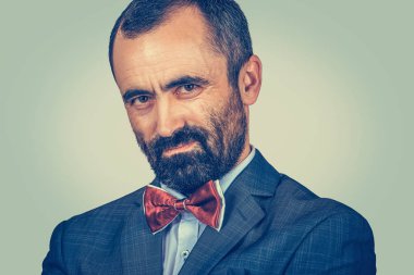 Kameraya yakın plan sakallı adam suçlayıcı görünüyor. İnsani duygu yüz ifadesi, vücut dili. Takım elbiseli, beyaz gömlekli, kırmızı papyonlu bir iş adamı. Tek bir kişi. duvar arkaplanı.