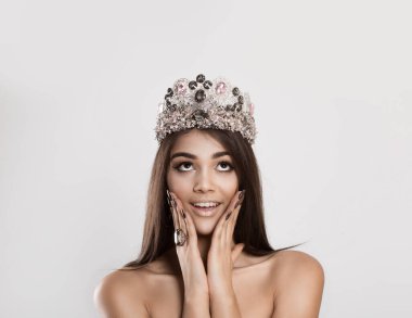 Wow, look up here. Happy beauty queen indian woman with crown looks up excited surprised, hands on her cheeks, against a white gray background