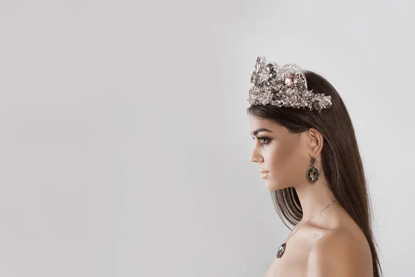 Closeup portrait in profile Beauty queen pageant winner girl with tiara long hair lipstick and eye shadows matching with dark crystal diamond earring necklace and crown looking to side on white grey — 스톡 사진