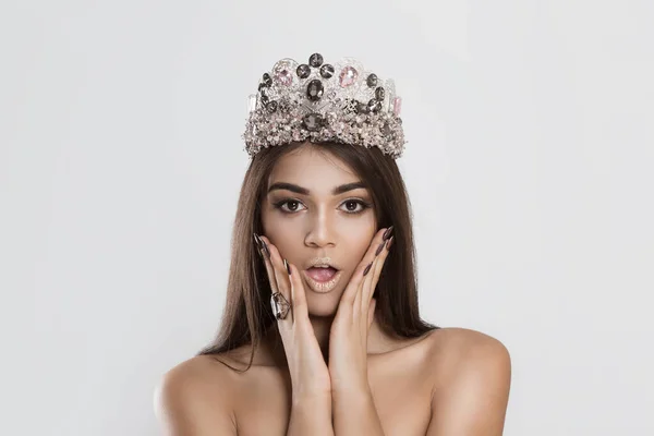 Wow, I won. Closeup young woman beautiful girl crowned miss long dark hair looking excited holding her mouth opened hands on cheeks white grey wall. Shocked surprised stunned. Positive human emotion