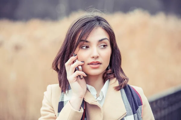 Woman on a telephone conversation. Girl in autumn beige coat blue white scarf speaking on mobile phone isolated outdoors light brown background