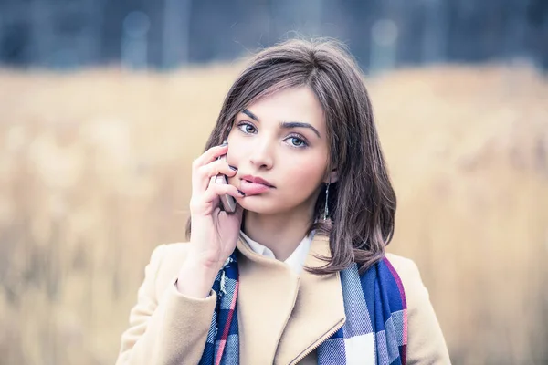 Woman girl talking on a mobile phone playing with her lips looking at you camera isolated outdoors light brown park background in autumn nature, wearing beige coat, blue white scarf — Stok fotoğraf