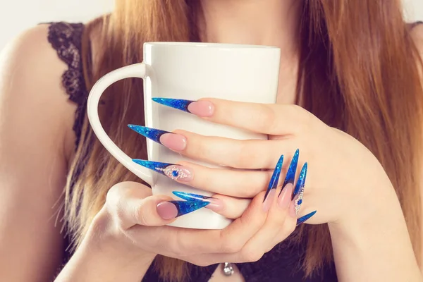 Nail Polish. Modern style new trend blue stilletto Nail Polish. Beauty hands holding white cup with hot beverage tea coffee. Stylish Colorful stiletto Nails isolated white background.  Art Manicure.