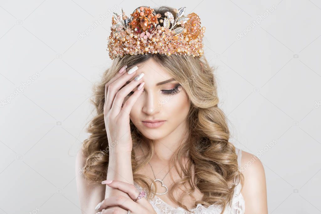 Sad Beautiful blonde girl in a golden crown on white grey background, eyes closed looking down with grief hand on shoulder pink ring on it