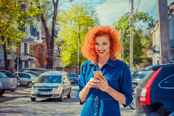 Aplicação de táxi. Mulher urbana sorridente muito feliz usando telefone inteligente, de pé na rua da cidade no dia ensolarado à espera de táxi. Menina ruiva bonita segurando móvel, mensagem SMS sms vestindo camisa jeans — Fotografia de Stock