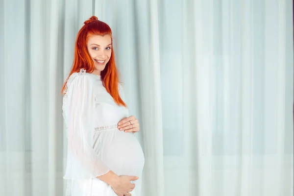 Enceinte. Portrait de jeune rousse enceinte de 39 semaines jolie femme souriante heureuse debout près de la fenêtre rideau blanc habillé de vêtements de robe blanche décontractée — Photo