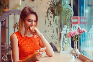 Yumruk ısırmak ve cep telefonuna bakmak. Kahve dükkanında cep telefonuyla kızgın bir kızın yan görüntüsü, pencerenin yanındaki evde oturma odası.