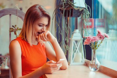 Yumruk ısırmak ve cep telefonuna bakmak. Kafede ya da pencerenin yanındaki evde cep telefonuyla kızgın bir kızın yan görüntüsü.
