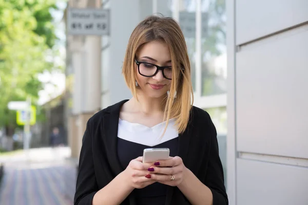 Singola signora felice utilizzando uno smartphone guardando on line contenuti in piedi sulla strada sorridente in abiti da lavoro e occhiali neri . — Foto Stock