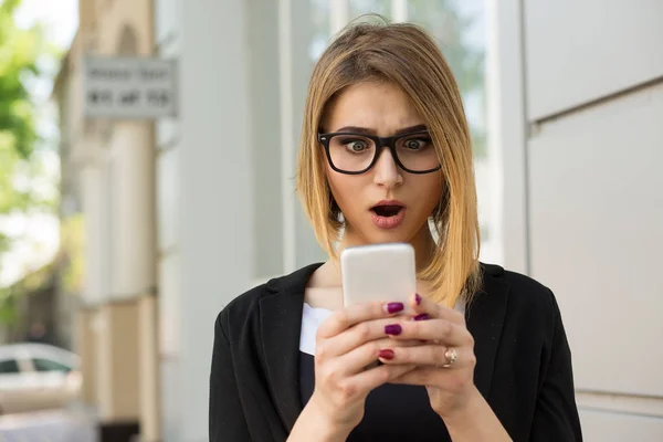 Donna scioccata con sms messaggio al telefono. Primo piano ragazza donna guardando il cellulare stordito volto sorpreso, abito nero abbigliamento formale, occhiali da vista, bob acconciatura al di fuori della città urbano home office su sfondo — Foto Stock