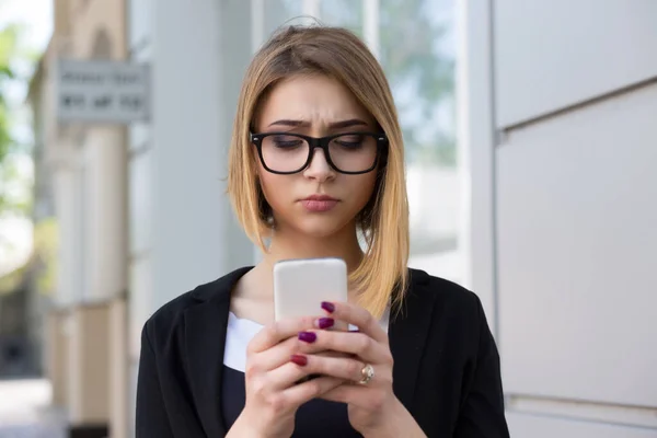 Un messaggio sconvolgente. Vista frontale di una triste ragazza depressa che controlla un messaggio telefonico che cammina per strada. Modello di razza mista in abiti da lavoro neri e occhiali — Foto Stock