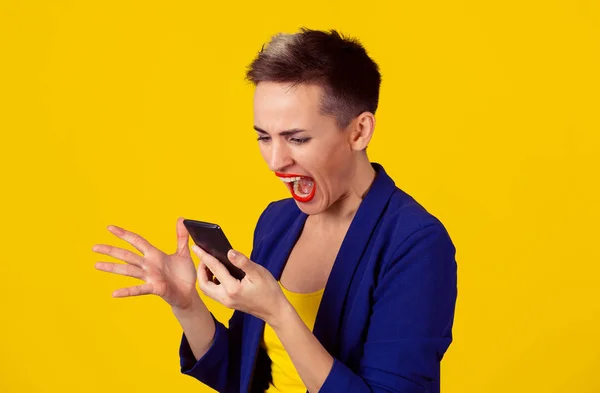 Retrato mujer joven enojada gritando mirando en el teléfono móvil aislado en el fondo amarillo de la pared. Emociones humanas negativas sentimientos. Pelo corto, traje azul, camisa amarilla — Foto de Stock