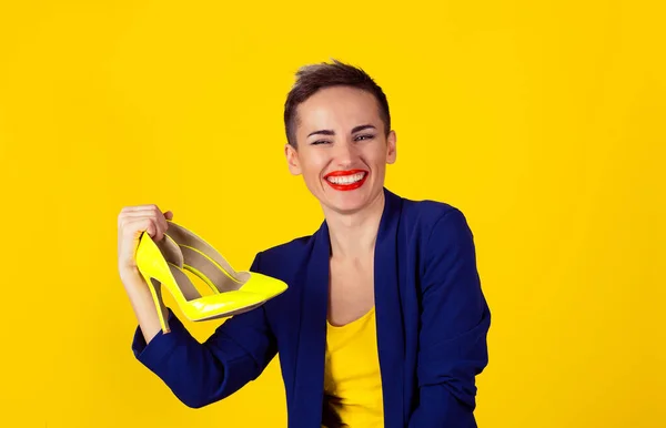 Mulher feliz sorriso dos dentes mostrando seus sapatos de salto alto. Multicultural latina menina cabelo curto lábios vermelhos azul terno camisa amarela isolada parede de fundo amarelo com espaço de cópia — Fotografia de Stock