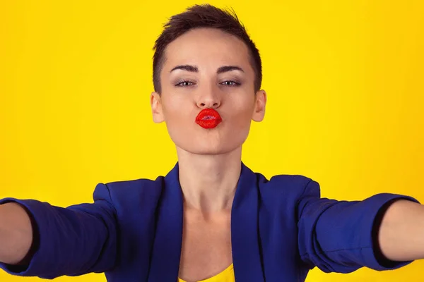 Öpüşme, selfie çekimi. Yakından kısa saçlı bir kadın sanal bir öpücük için dudaklarını büzüştürürken cep telefonu kamerasında selfie çekiyor ve izole edilmiş sarı arka planı soruyor. — Stok fotoğraf