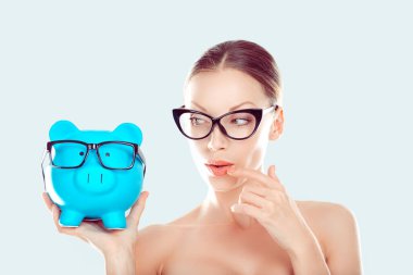 Kapanış stresi çeken genç kadın domuz kumbarasına bakıyor, düşünüyor, finansal planlar üzerinde kafa yoruyor açık mavi arka plan. Ekonomik borç kavramı para zorlukları. Olumsuz duygu ifadesi