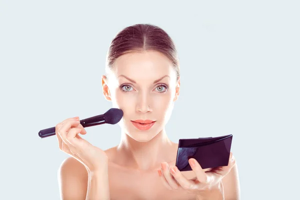 Make up. Close up portrait  head shot  young woman pretty smiling girl teenager applying foundation powder, blusher with brush holding mirror looking at you camera isolated light blue white background — Stock Photo, Image