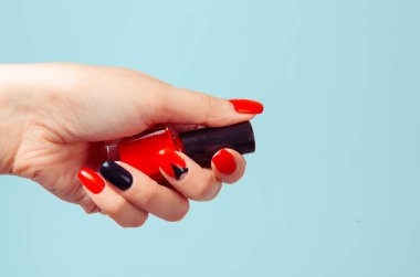 Elinde oje şişesi tutan bir kadın. Sanat manikürü. Modern stil kırmızı siyah ojeli üçgen. Renkli topuklu çiviler izole edilmiş mavi arka plan güzellik manikürlü güzel kadın eli