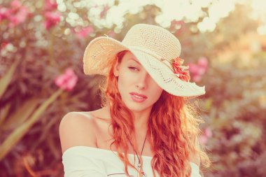 Hasır şapkalı, beyaz dişli bir kadın yazın parkta düşünür ve yan bakar, arka planda çiçek çalısı