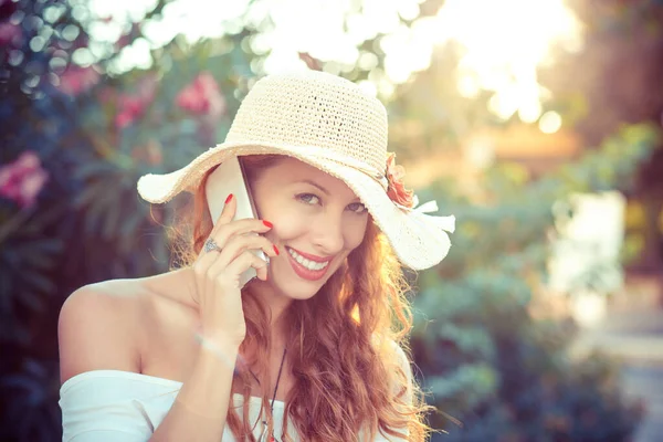 Ευτυχισμένος στο κινητό τηλέφωνο. Ευφορική γυναίκα με ψάθινο καπέλο που μιλάει στο έξυπνο κινητό της πράσινο θάμνο στο φόντο στο ηλιοβασίλεμα — Φωτογραφία Αρχείου