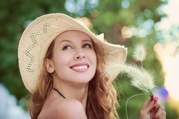 Happy outside. Cheerful beautiful girl in straw hat looking up smiling holding green herb flower in sunny summer park — Stock Photo, Image