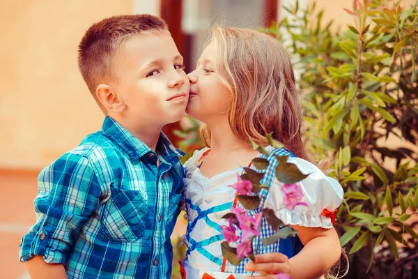 Irmã está beijando seu irmão na bochecha, abraços quentes na natureza fora perto da casa. Ele parece e sorri feliz. Menina mostra carinho para seu amigo menino, beijando-o na bochecha — Fotografia de Stock