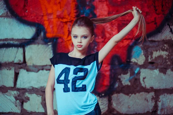 Petite fashionista. Portrait petite fille enfant posant tirant sur sa longue queue de cheval blonde cheveux graffiti isolé fond de brique blanche, chemise bleu clair avec le numéro 42 dessus, rouge cassé entendu sur le mur — Photo