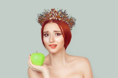Güzellik kraliçesi vitamin şekeri için can atıyor. Kafasında kristal taç olan güzel bir kadın elinde yeşil elmayla açık yeşil arka plan duvarında izole edilmiş dudaklarını ısırıyor..