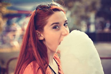 Kızıl saçlı kız kameraya bakıyor. Açık havada pamuk şeker yiyor. Arka planda, eski moda.. 