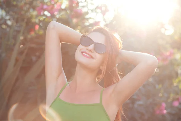 Primer plano retrato feliz joven bonita mujer en camisa verde con gafas de sol negras mirando hacia arriba, las manos en la cabeza en el aire relajante, arbusto verde con flores de color rosa en el fondo. Técnicas de alivio del estrés — Foto de Stock