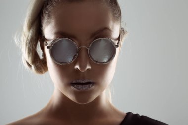 Gri metalik aynalı, parlak güneş gözlüklü bir kadın. İleride kamerana bakıyor. Saçları at kuyruğu örgülü beyaz arka plan.