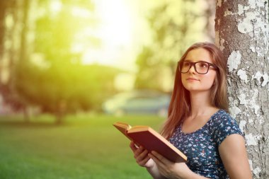 Roman hakkında düşünüyorum. Gün batımında bir ağacın yanında kağıt kitap okuyan mutlu bir kadının yan görünüm portresi. Karışık Asyalı Rus ırkı modeli