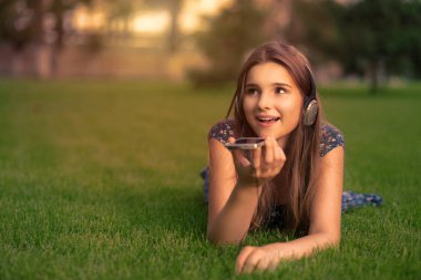 Öğrenci, kafasında kulaklıkla parkta çimlerin üzerinde oturan notları kaydetmek için akıllı bir telefonla ses tanıma sistemi kullanıyor.