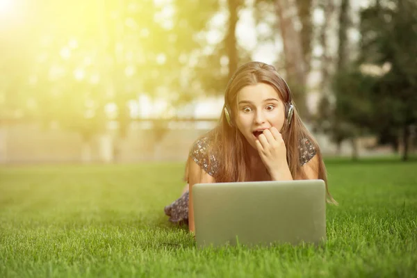Buenas noticias. Mujer emocionada asombrada sorprendida por lo que ve en su computadora portátil tumbada en el césped de hierba verde afuera en los auriculares del parque de la ciudad en la cabeza — Foto de Stock