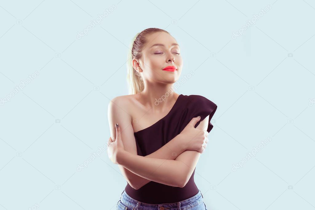 Love yourself concept. Smiling woman holding hugging herself isolated light blue wall background. Positive human emotion, facial expression.