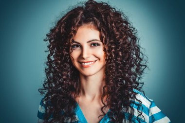 Mavi arka planda izole edilmiş kameraya bakarak gülümseyen neşeli, genç, kıvırcık bir kadının yakın portresi.