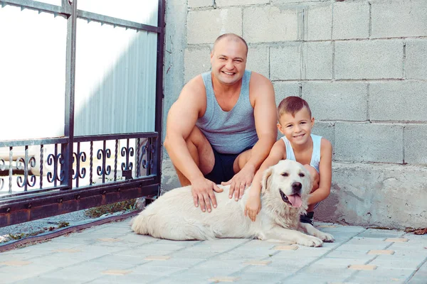 Father And Son Sitting playing posing With Dog outside the house smiling near fence bricks wall background