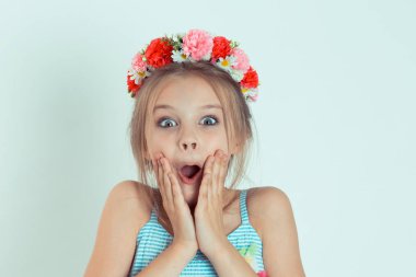 Vay canına. Güzel kız ağzını açık tutarak heyecanlanıyor, elleri yanaklarında. Şok oldum. Olumlu duygular. Açık mavi arka planda çiçekli saç bandıyla beyaz çocuk modelinin yakın plan portresi